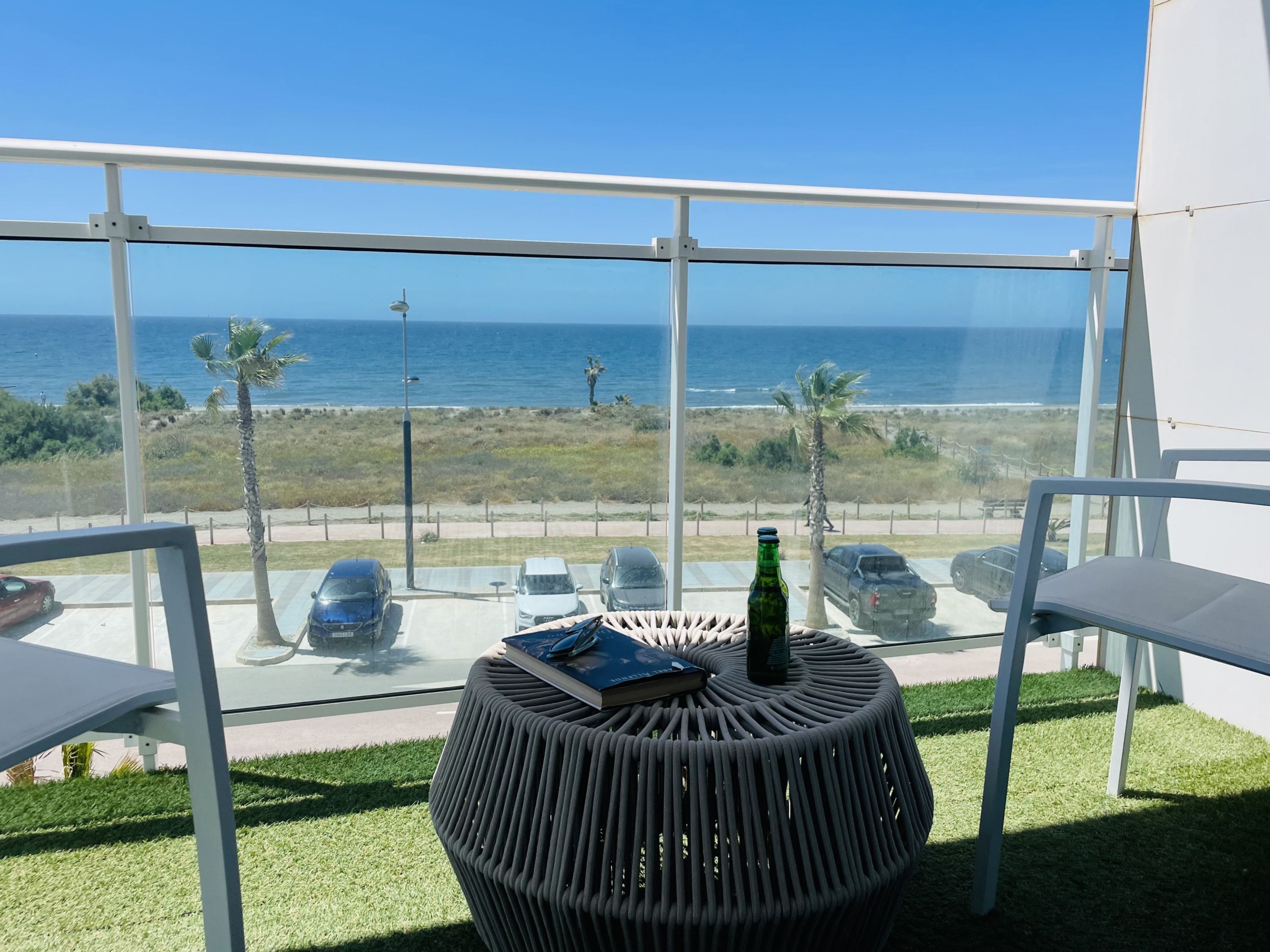 A balcony with a view of the ocean, a round table with a book and a bottle, and a chair.