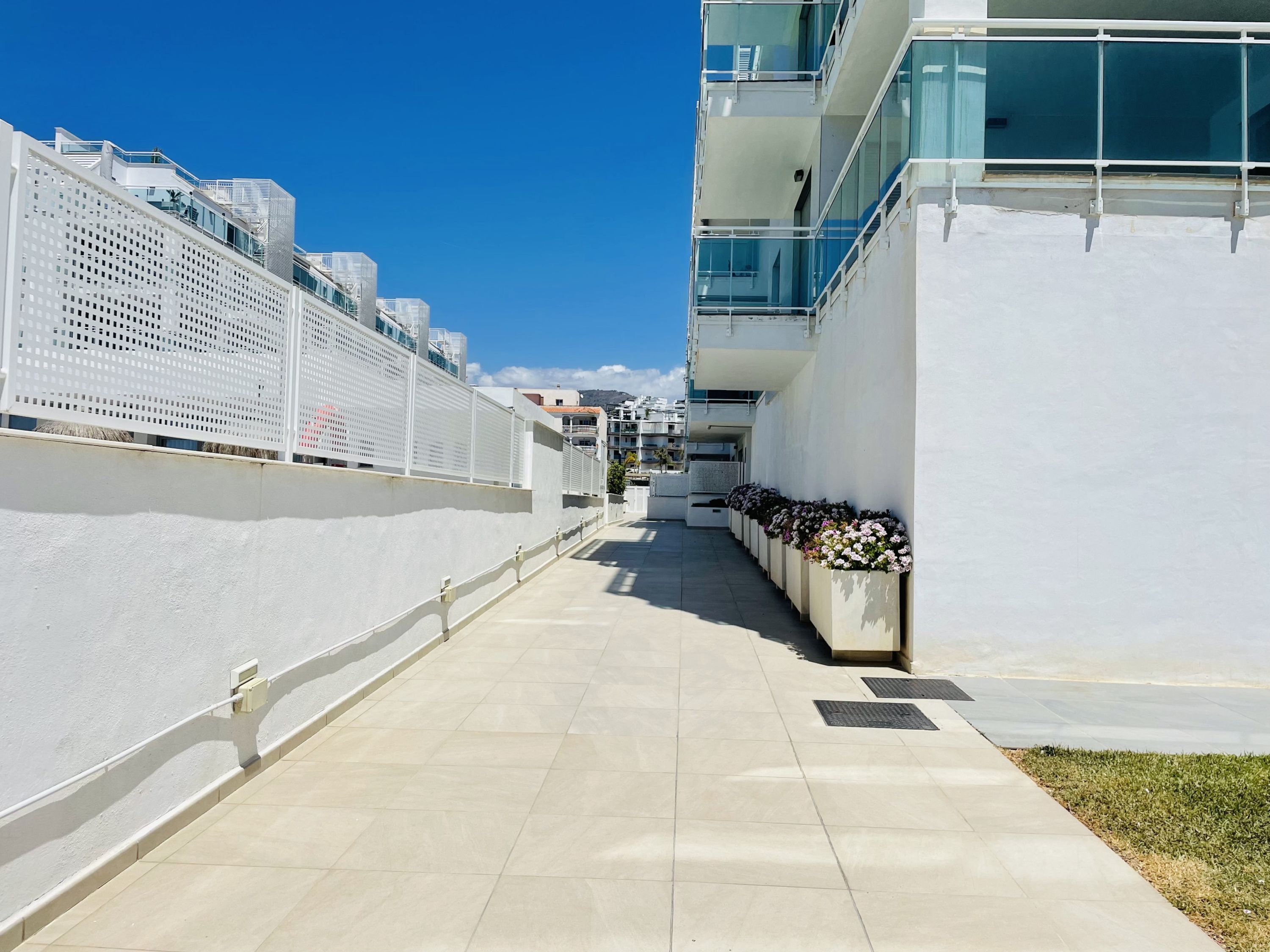 Un edificio moderno con un largo pasillo recto bordeado de macetas y una cerca blanca a un lado.