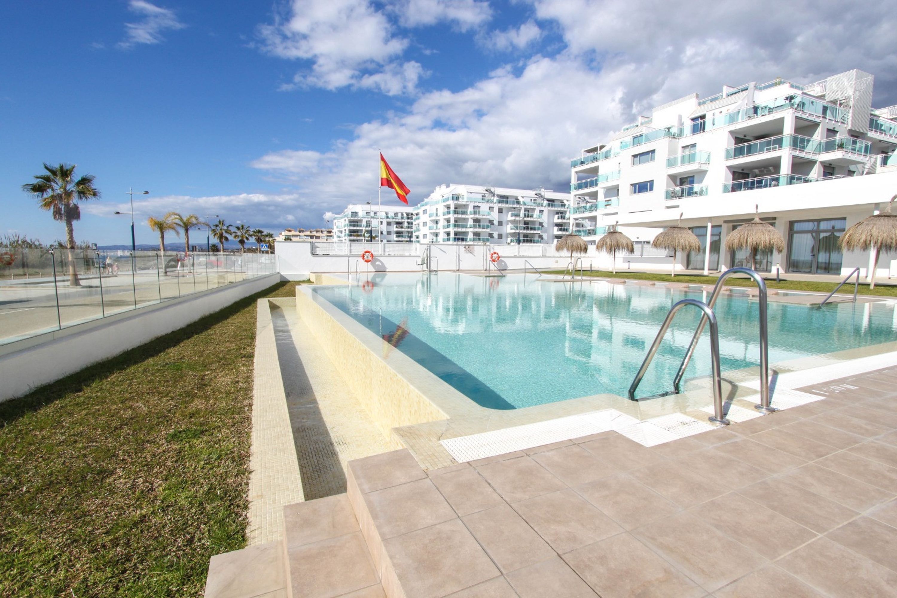 A swimming pool with a clear view of the sea, a flag, and a modern building with balconies.