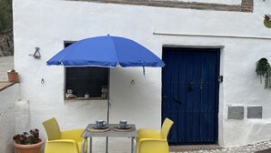 Um pátio com uma mesa, duas cadeiras amarelas, um guarda-sol azul, uma porta azul e plantas em vasos.