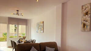 A living room with a brown sofa, two patterned cushions, a dining table with chairs, a chandelier, and a large window with blinds. - Apartamento Torrox Costa a Metros de la Playa (Torrox)
