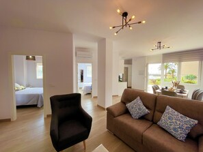Una sala de estar moderna con un sofá marrón y un sillón negro, un comedor con mesa y sillas, y una cocina al fondo.