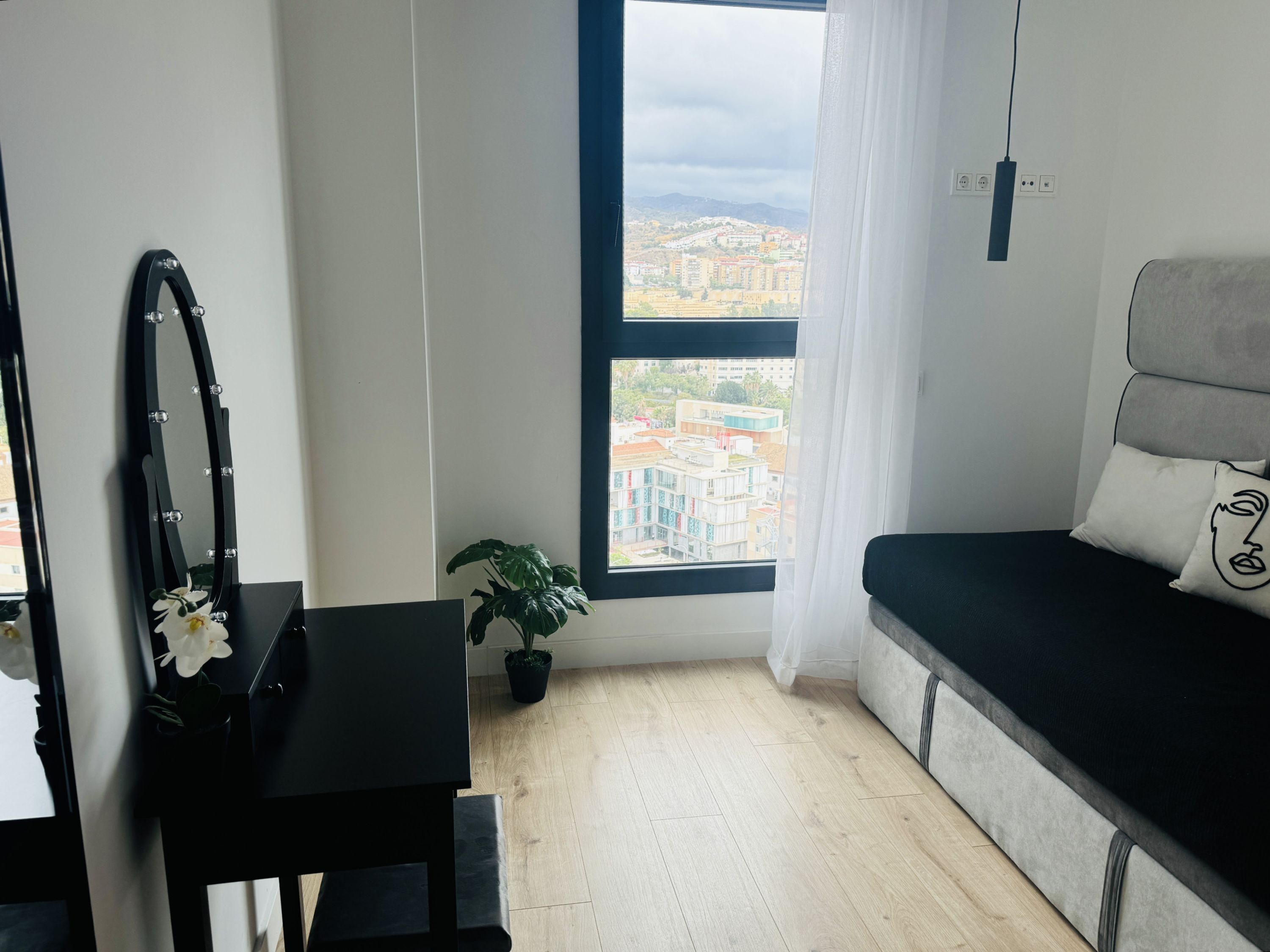 Une chambre moderne dotée d’une grande fenêtre, d’un canapé noir, d’une petite table et d’un miroir avec éclairage intégré.