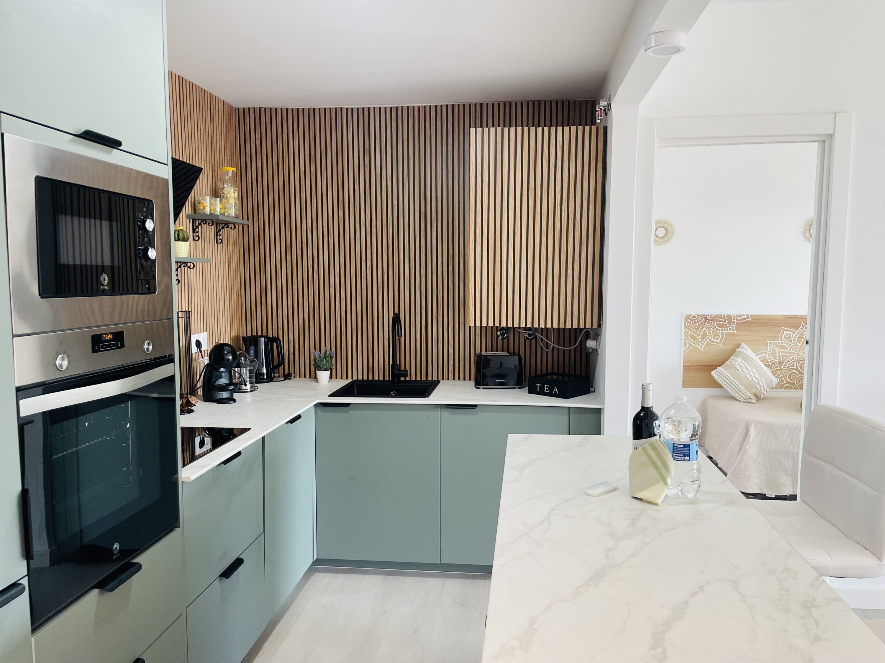 A modern kitchen with a built-in oven, microwave, and coffee maker. There is a sink, a countertop with a tea box, and a bottle of water. The kitchen has a wooden panel wall and a white countertop.