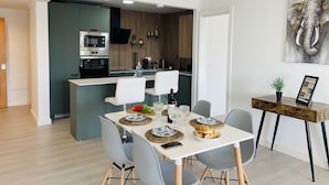 Una cocina moderna con electrodomésticos integrados, un comedor con mesa blanca y sillas grises, y una sala de estar con una mesita de madera y una planta.