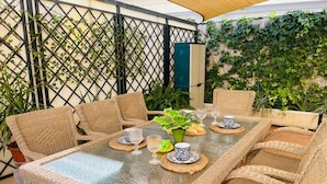 A covered outdoor dining area with wicker furniture and a glass table, surrounded by greenery and a bamboo fence. - Apartamento Cantarero by At Home Costa del sol (Nerja)