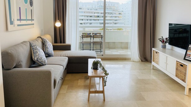 Un soggiorno moderno con un divano, un tavolino con una pianta, un televisore su un mobile e un balcone con vista sugli edifici.