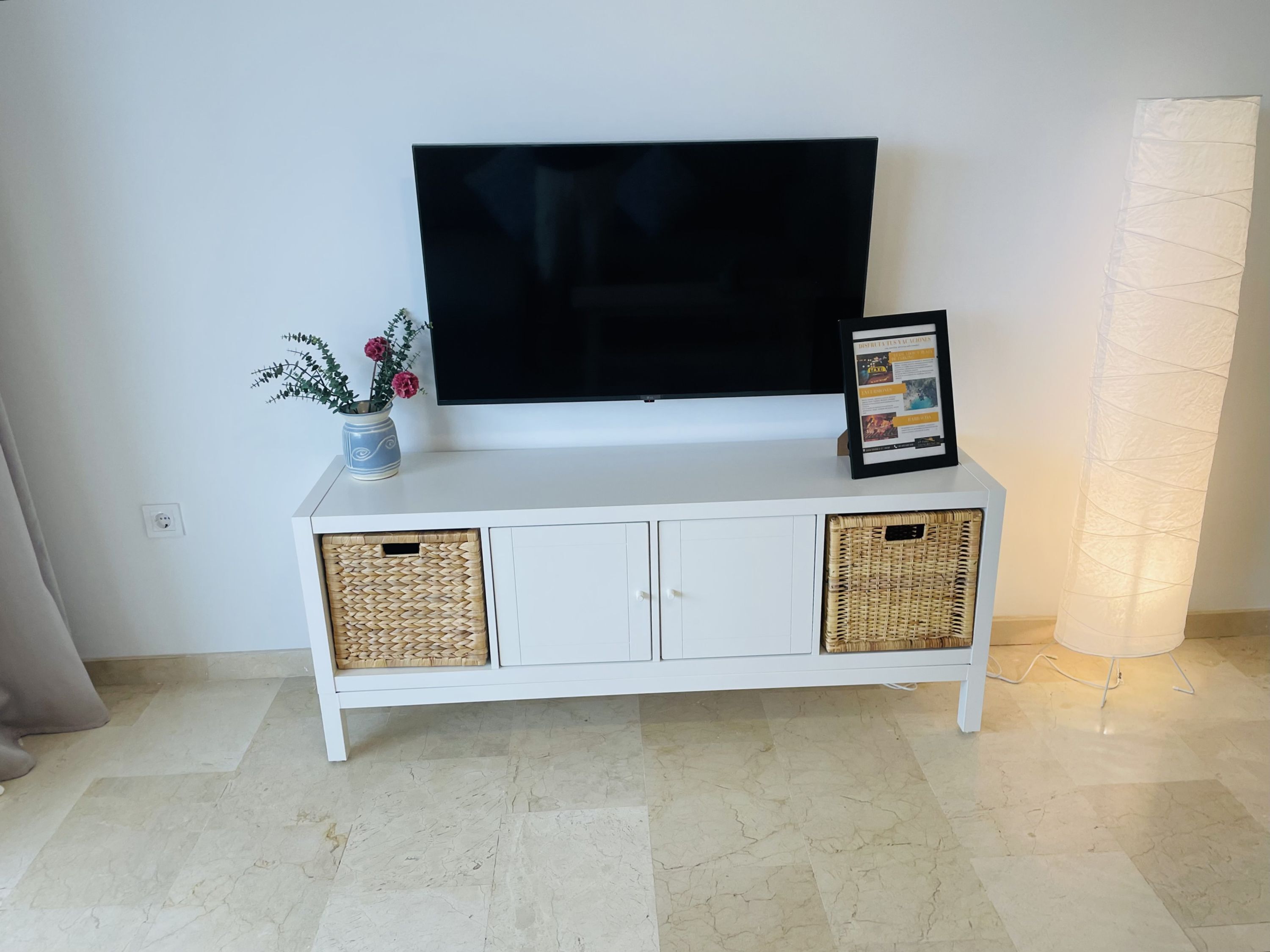 Un meuble de rangement blanc pour appareils multimédias, avec un téléviseur à écran plat, un tableau encadré et un vase avec des fleurs.