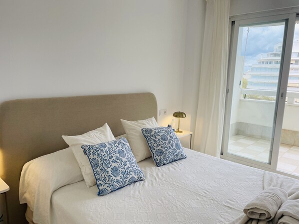 A hotel room with a bed, two pillows, a nightstand, a lamp, and a view of the outside. - Apartamento Sol de Marbella Sea View by At Home (Marbella)