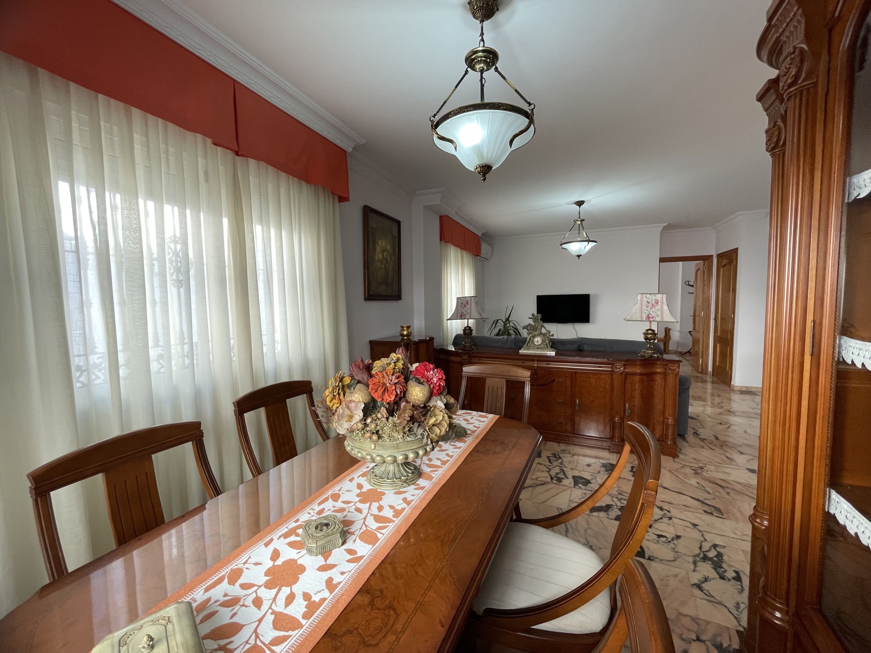 A dining area with a long wooden table, chairs, a vase with flowers, and a television in the background.