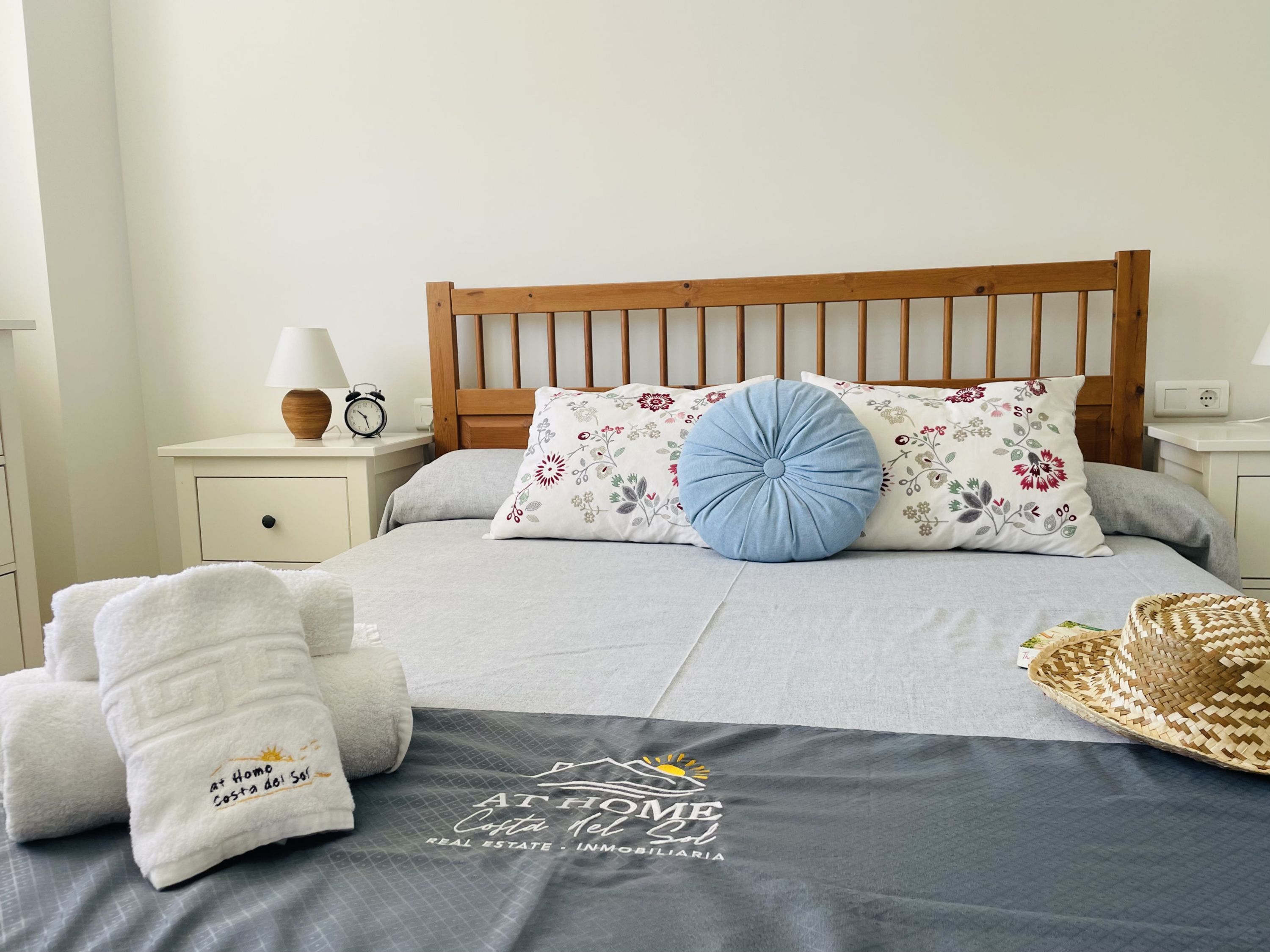 Een net opgemaakt bed met een grijze deken, twee kussens met bloem- en blauwe motieven, een houten hoofdbord, een nachtkastje met een lamp en een wekker, en een geweven mandje op het bed.