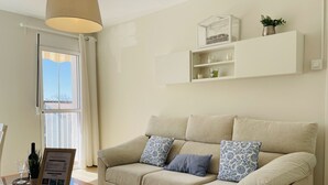 Una sala de estar con un sofá color beige, una mesa de centro de cristal, un estante blanco montado en la pared con objetos decorativos, y una ventana con cortinas.
