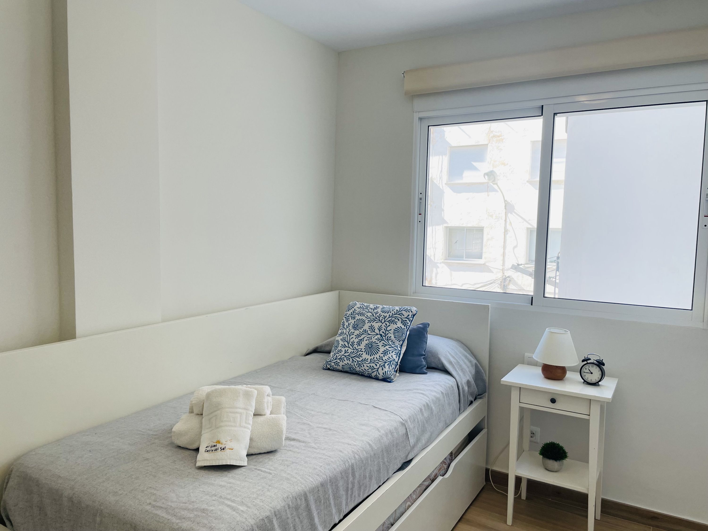 Quarto individual com janela, mesa de cabeceira com abajur e despertador, e uma pilha de toalhas.