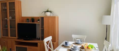 Ein modernes Wohnzimmer mit Fernseher, Holzschrank, Esstisch mit Stühlen und einer kleinen Küchenzeile.