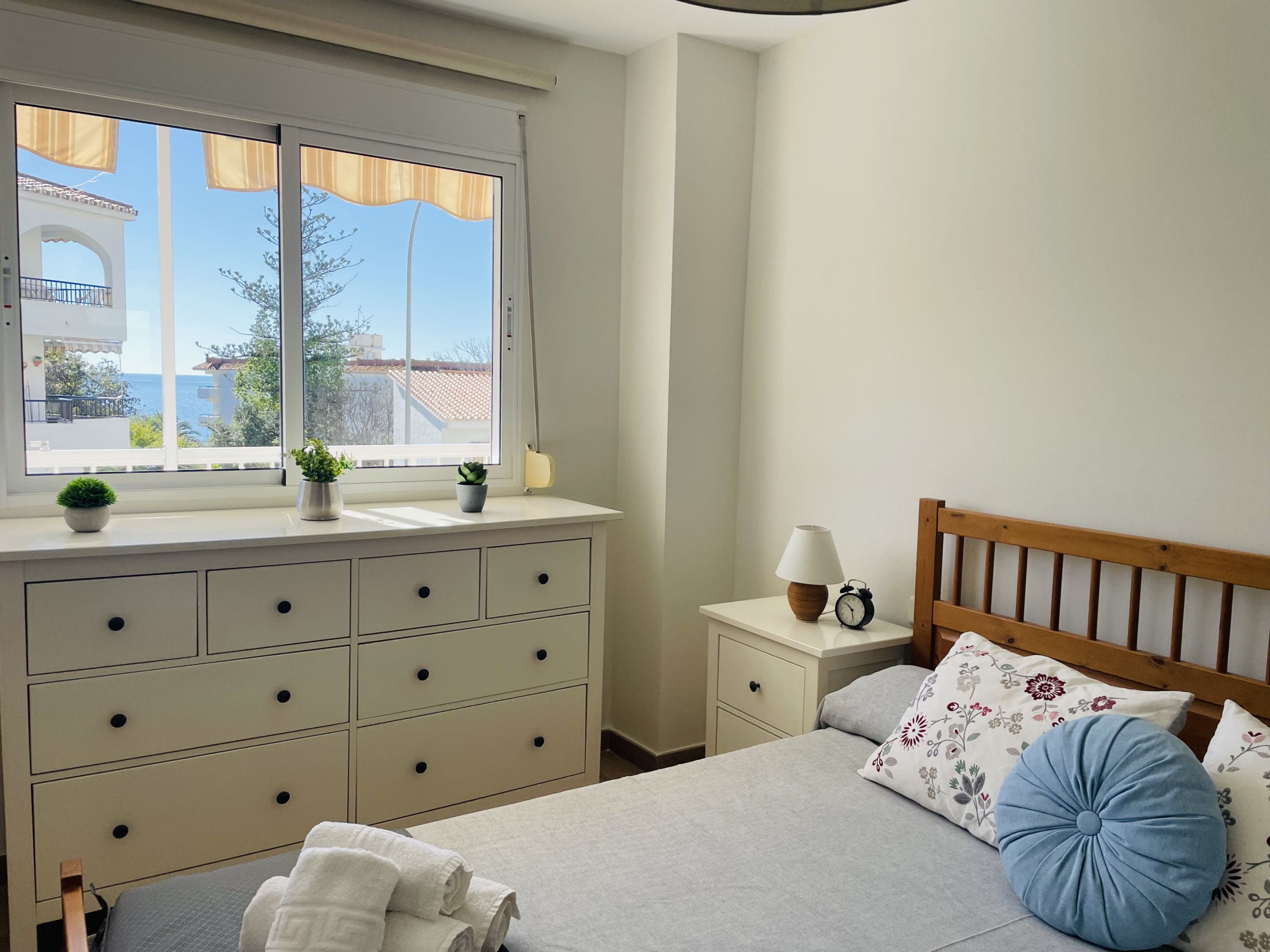 A bedroom with a bed, a nightstand, a dresser, a window with curtains, and a view of buildings and trees.