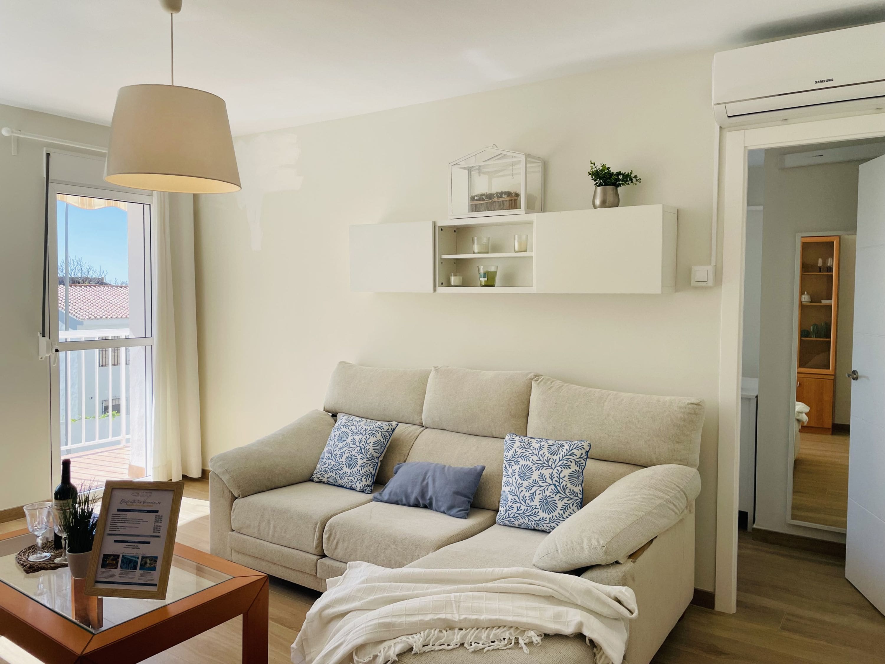 Ein Wohnzimmer mit einem beigen Sofa, einem Glastisch, einer Pendelleuchte und einem weißen Regal mit Kerzen.