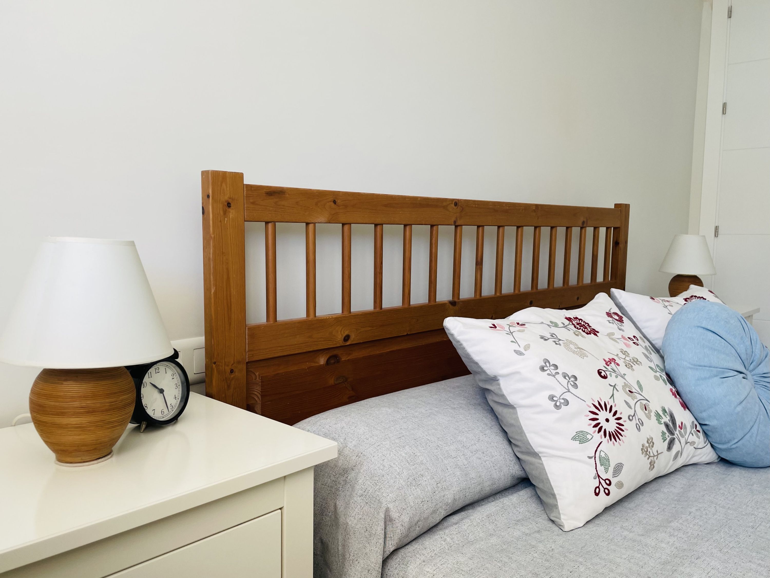 Une chambre avec une tête de lit en bois, une table de chevet blanche avec une horloge et une lampe, un lit gris avec des coussins à motifs floraux et un mur blanc.