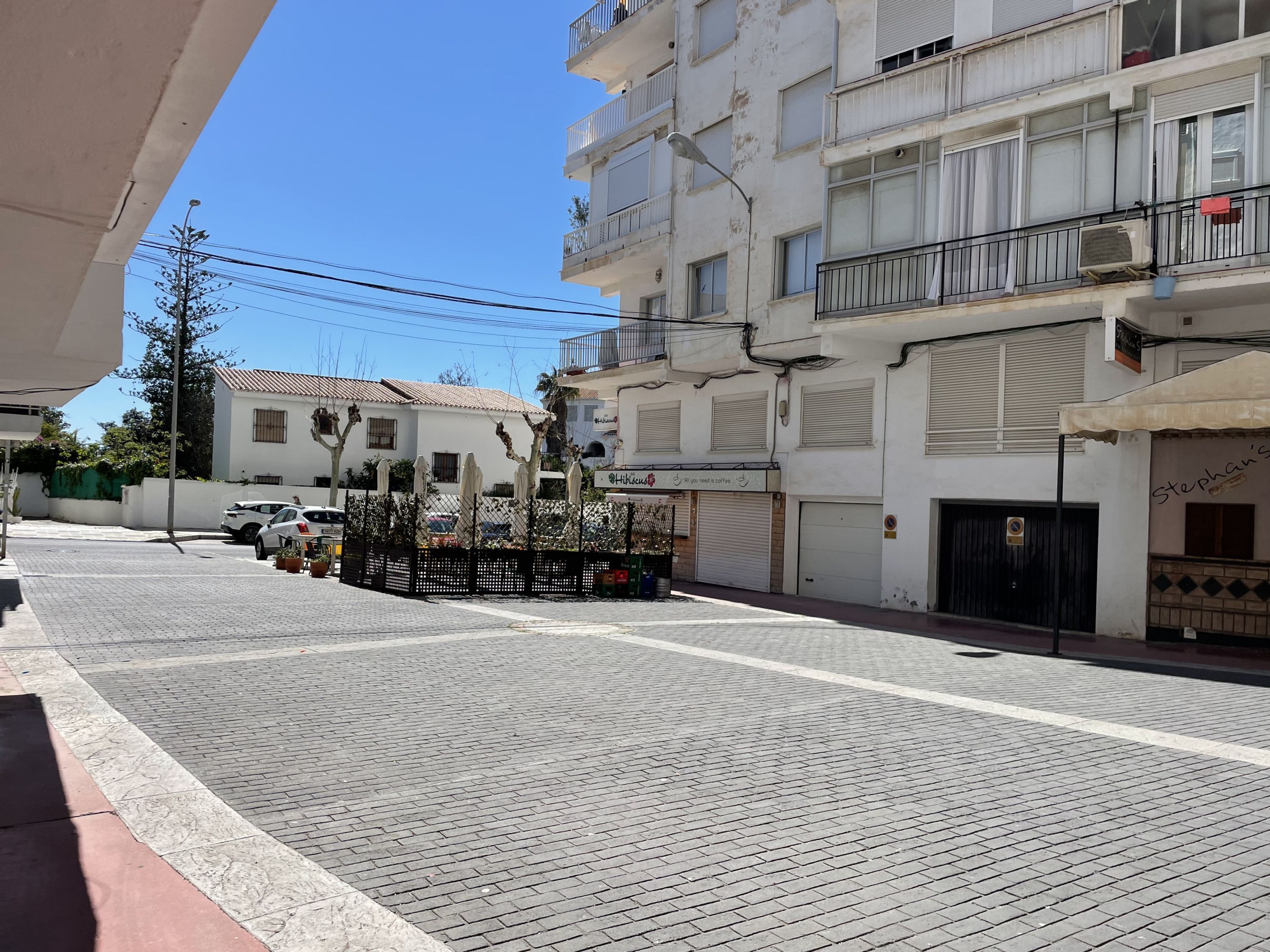 A street with a paved sidewalk, parked cars, and buildings with balconies and awnings.