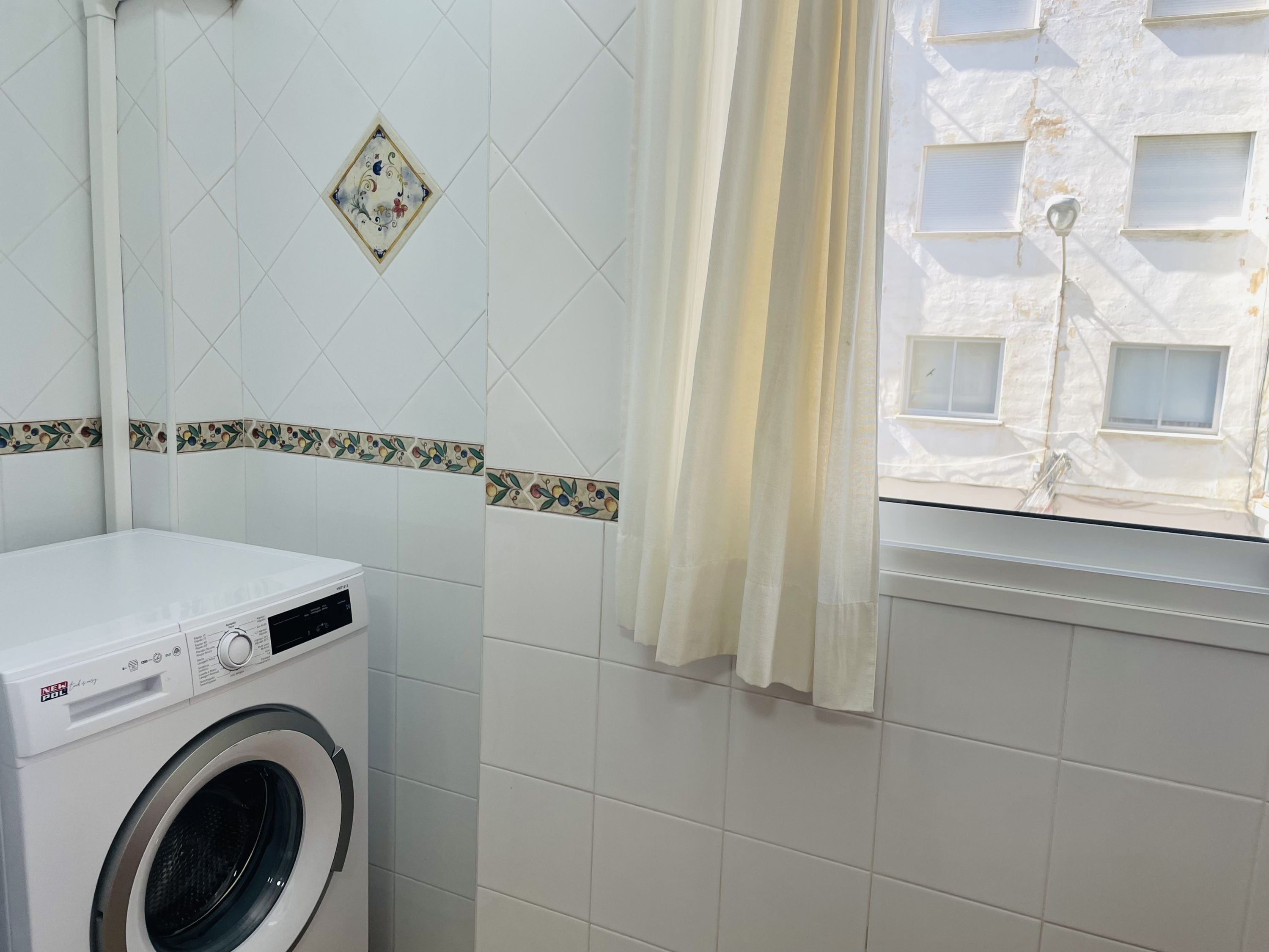 Een wasruimte met een wasmachine, een raam met gordijnen, en een betegelde muur met decoratieve rand.