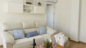 Una sala de estar con un sofá color beige, una mesa de centro de vidrio, una botella de vino y una planta en maceta.