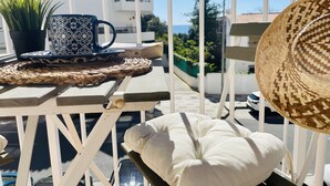 Un balcón con una mesa de mimbre, una planta en maceta, una taza estampada, un sombrero de paja y un cojín blanco.