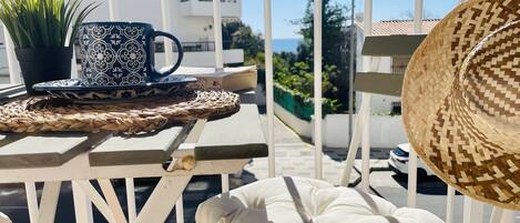 A balcony with a woven table, a potted plant, a patterned mug, a straw hat, and a white cushion.