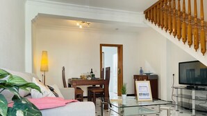 Una habitación de hotel moderna con una cama, una mesita de centro de vidrio, una zona de comedor con una mesa y sillas de madera, un televisor y una escalera con balaustres de madera.