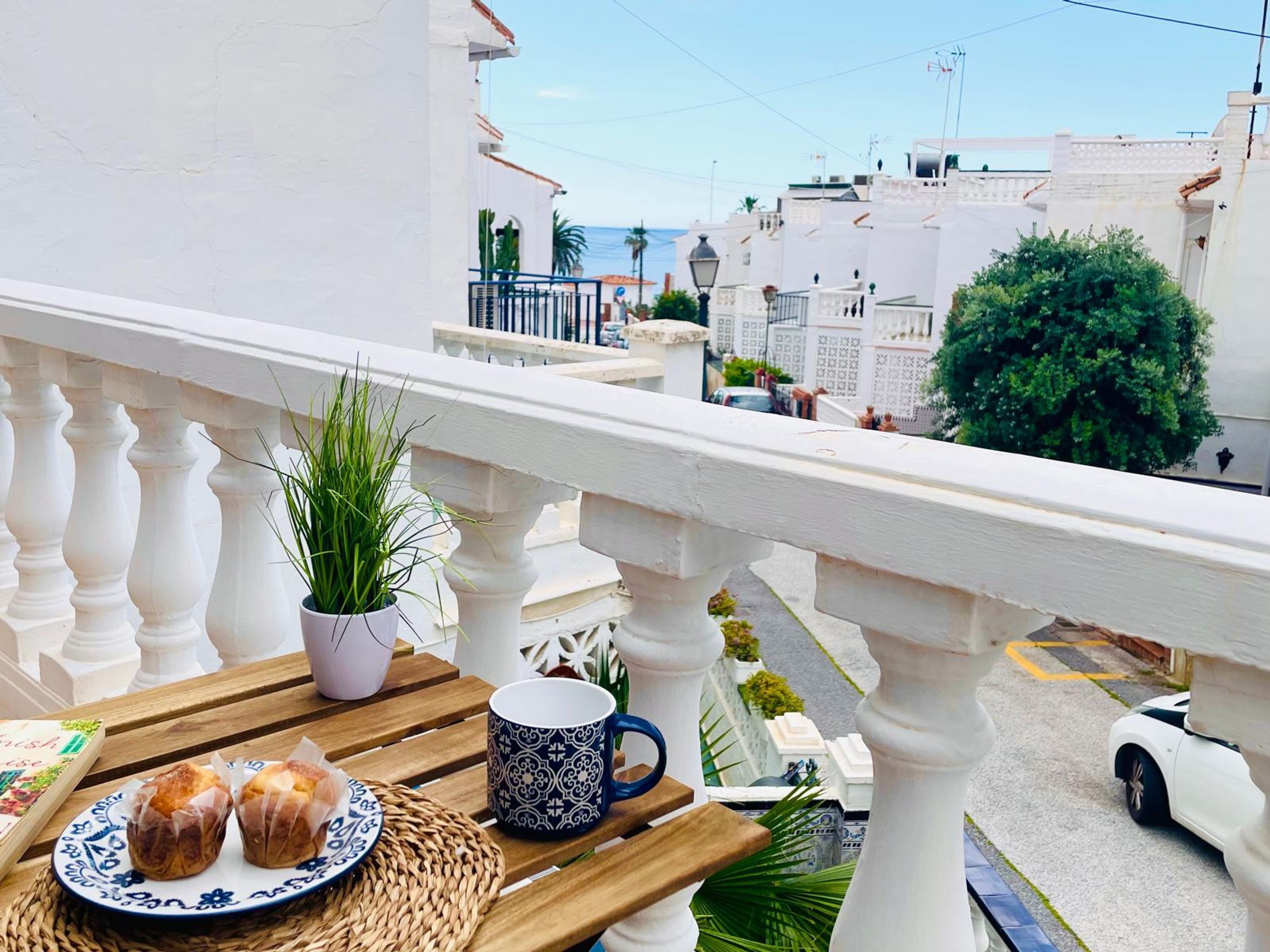 Varanda com uma mesa posta para o pequeno-almoço, incluindo pastelaria e uma chávena, com vista para uma rua com edifícios e um carro.