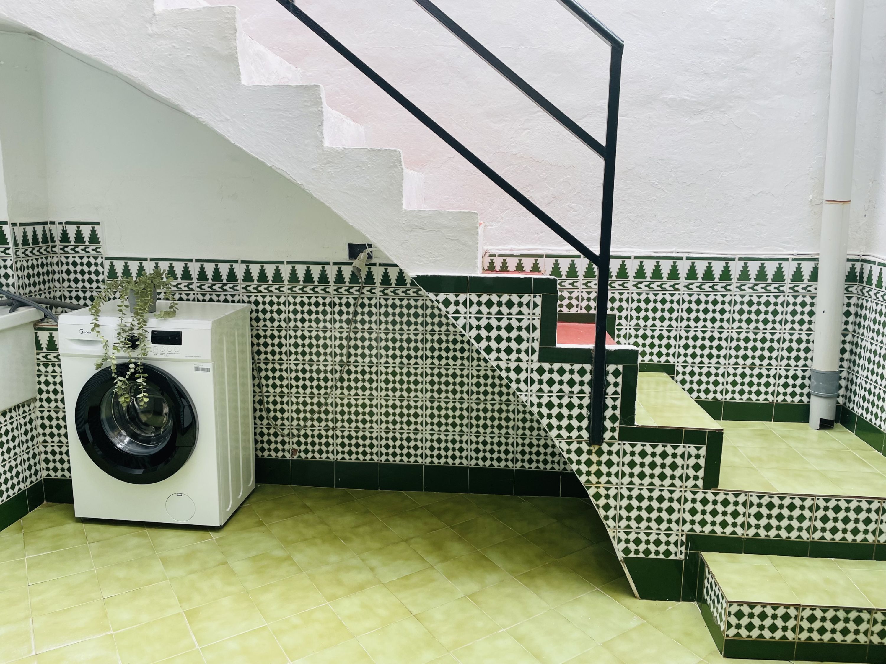 Ein Waschbereich mit Waschmaschine, eine Treppe mit grün-weißen gemusterten Fliesen und eine weiße Wand mit geometrischem Muster.