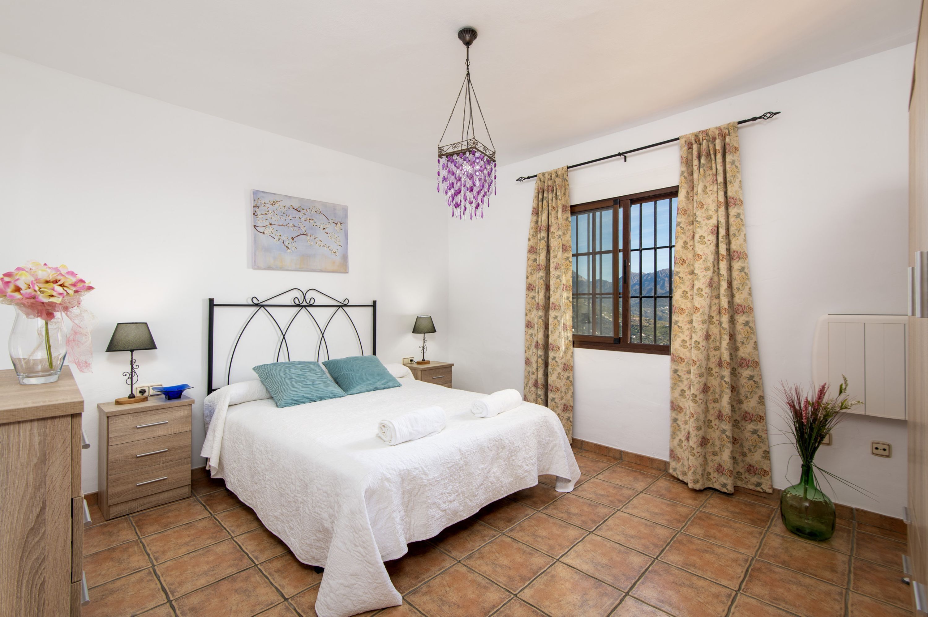 A bedroom with a bed, bedside tables, a nightstand, a window with curtains, a hanging light fixture, and a wall painting.