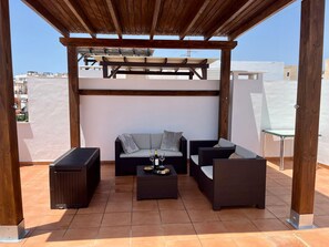 Una terrazza sul tetto con una pergola in legno, un divano imbottito, due poltrone, un tavolino con una bottiglia e dei bicchieri, e un cestino.