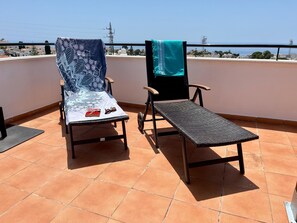 Zwei Liegestühle auf einer gefliesten Terrasse mit Blick auf das Meer und Gebäude in der Ferne.