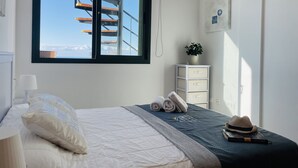 A bedroom with a bed, a window with a view of a spiral staircase, a bedside table with a plant, and a hat on the bed. - Apartamento Mar de Nerja Sunset (Nerja)