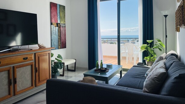 A living room with a large flat-screen TV, a sofa, a coffee table, and a view of the sea through a sliding glass door. - Apartamento Mar de Nerja Sunset (Nerja)