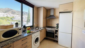 A compact kitchen with a built-in washer, a stainless steel sink, and a white refrigerator. A window offers a view of a hillside town. - Apartamento Mar de Nerja Sunset (Nerja)