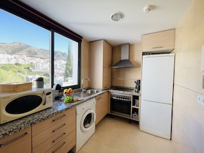 A compact kitchen with a built-in washer, a stainless steel sink, and a white refrigerator. A window offers a view of a hillside town. - Apartamento Mar de Nerja Sunset (Nerja)