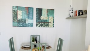 A dining area with a white table, two chairs, a wine bottle, and a plate of food. There are two abstract paintings on the wall above the table. - Apartamento Mar de Nerja Sunset (Nerja)