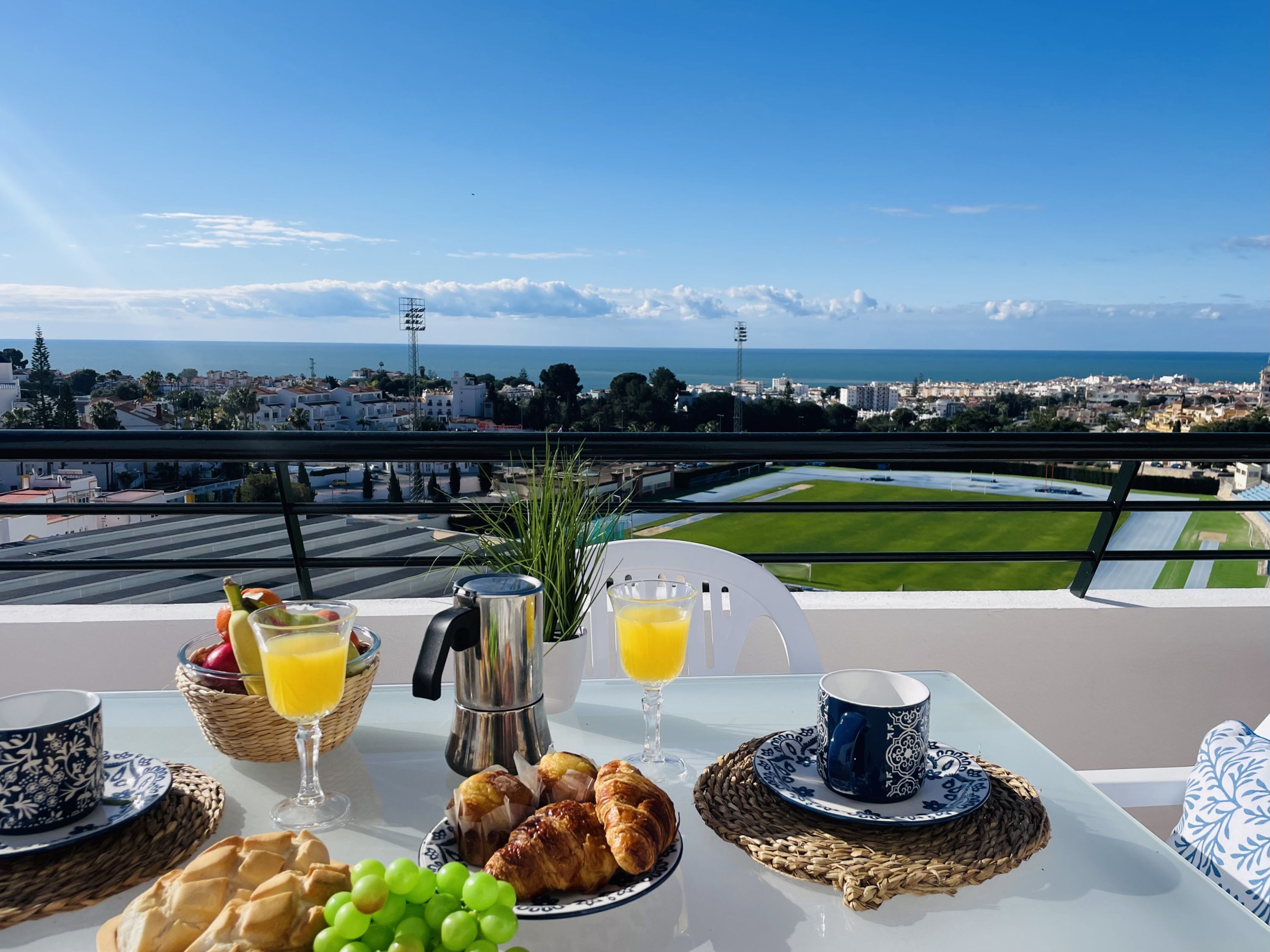 A breakfast table with croissants, grapes, and orange juice, overlooking a coastal cityscape with a clear sky.