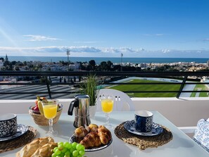 Une table de petit-déjeuner avec des croissants, des raisins et du jus d’orange, donnant sur un paysage urbain côtier sous un ciel dégagé.