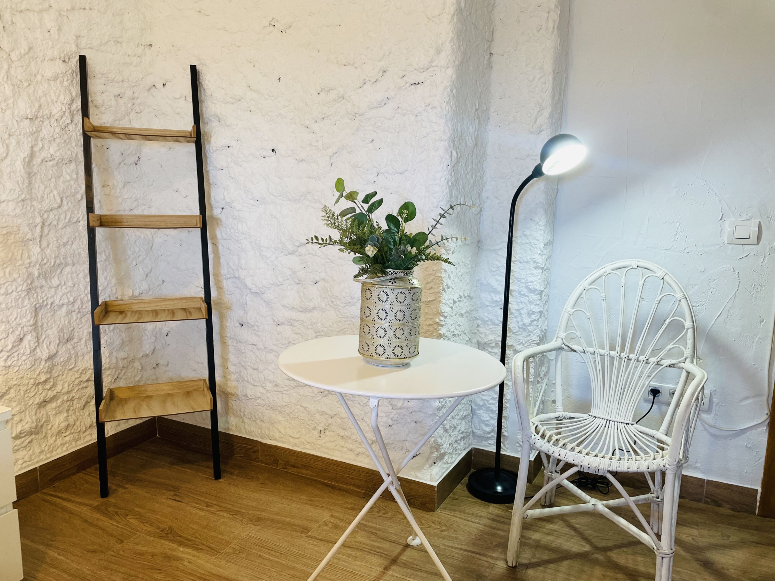 Une pièce d’angle avec un mur blanc à texture, une chaise en osier blanche, une petite table blanche sur laquelle est posée une plante en pot, un lampadaire et une échelle en bois.