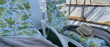 A balcony with two white plastic chairs covered with floral-patterned cushions, a grey towel, and a straw hat.