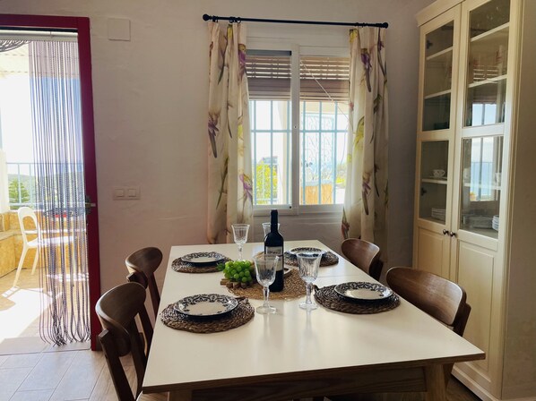 Una mesa de comedor dispuesta con platos, vasos y una botella, con vistas al exterior a través de una ventana con cortinas.