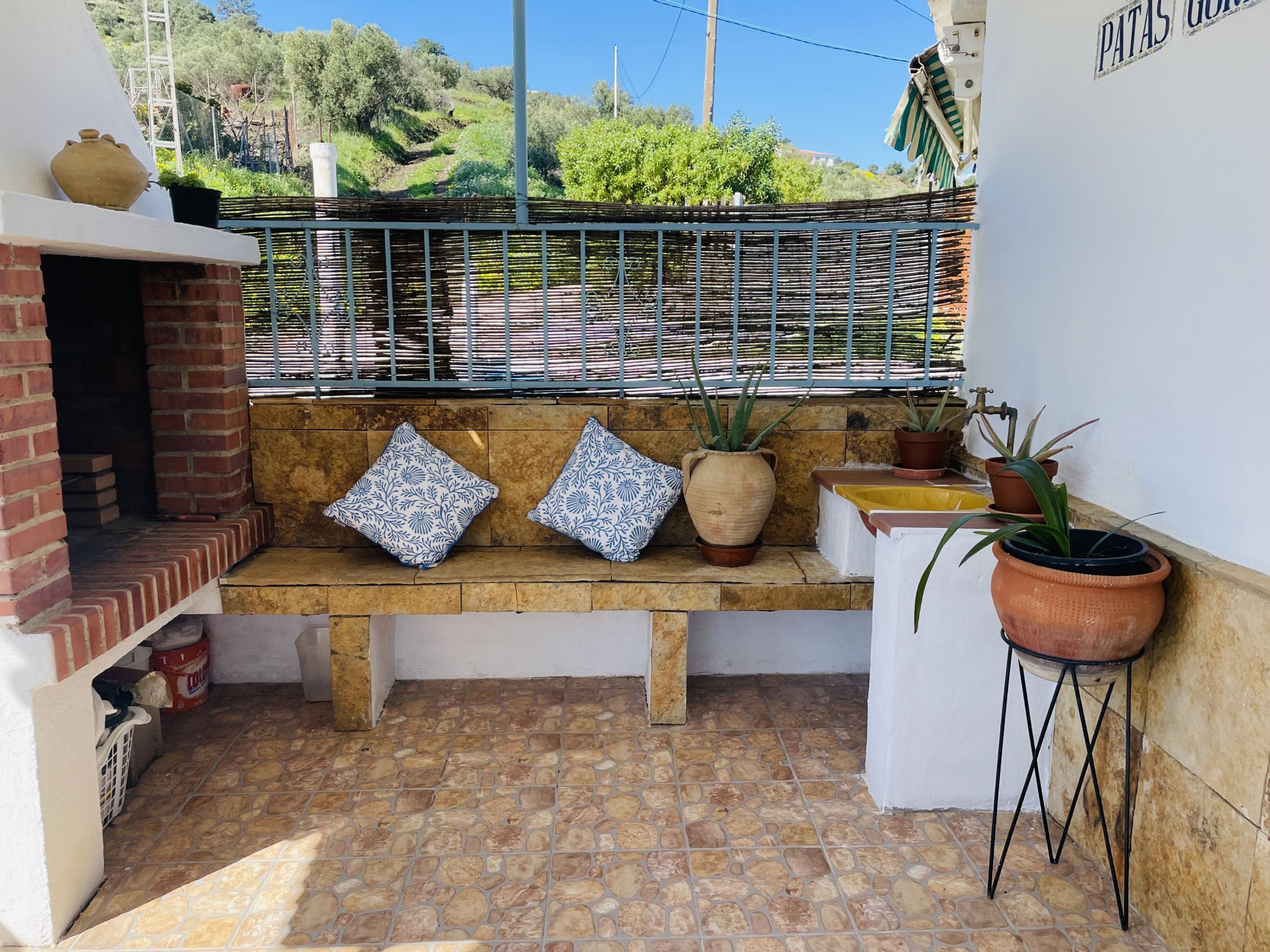 A patio area with a brick oven, a bench with patterned cushions, potted plants, and a metal fence.
