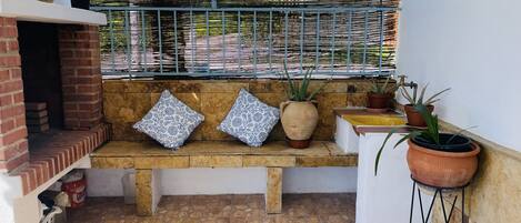 A patio area with a brick oven, a bench with patterned cushions, potted plants, and a metal fence.