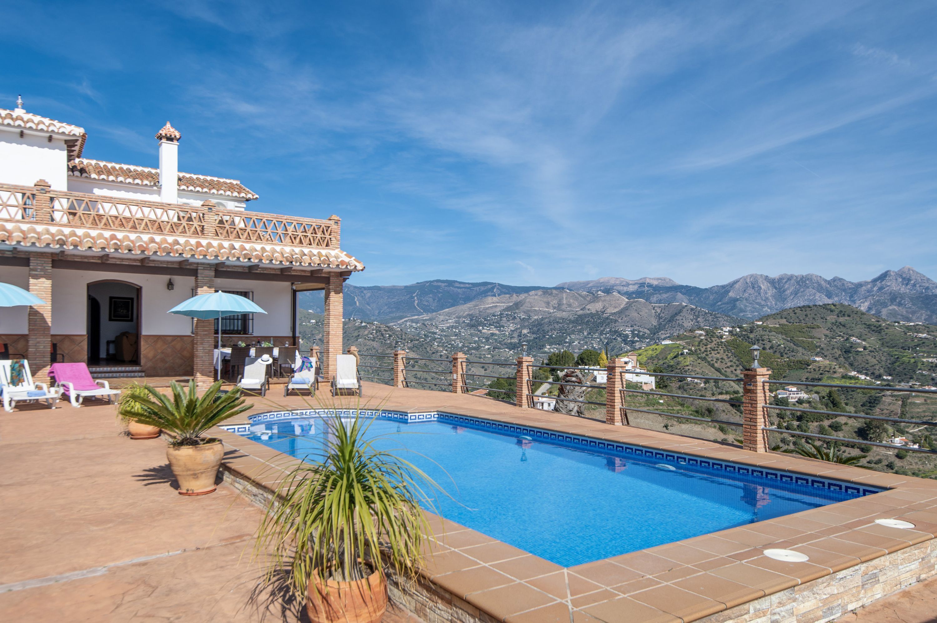 A swimming pool with a view of mountains, outdoor seating, and a building with a tiled roof.