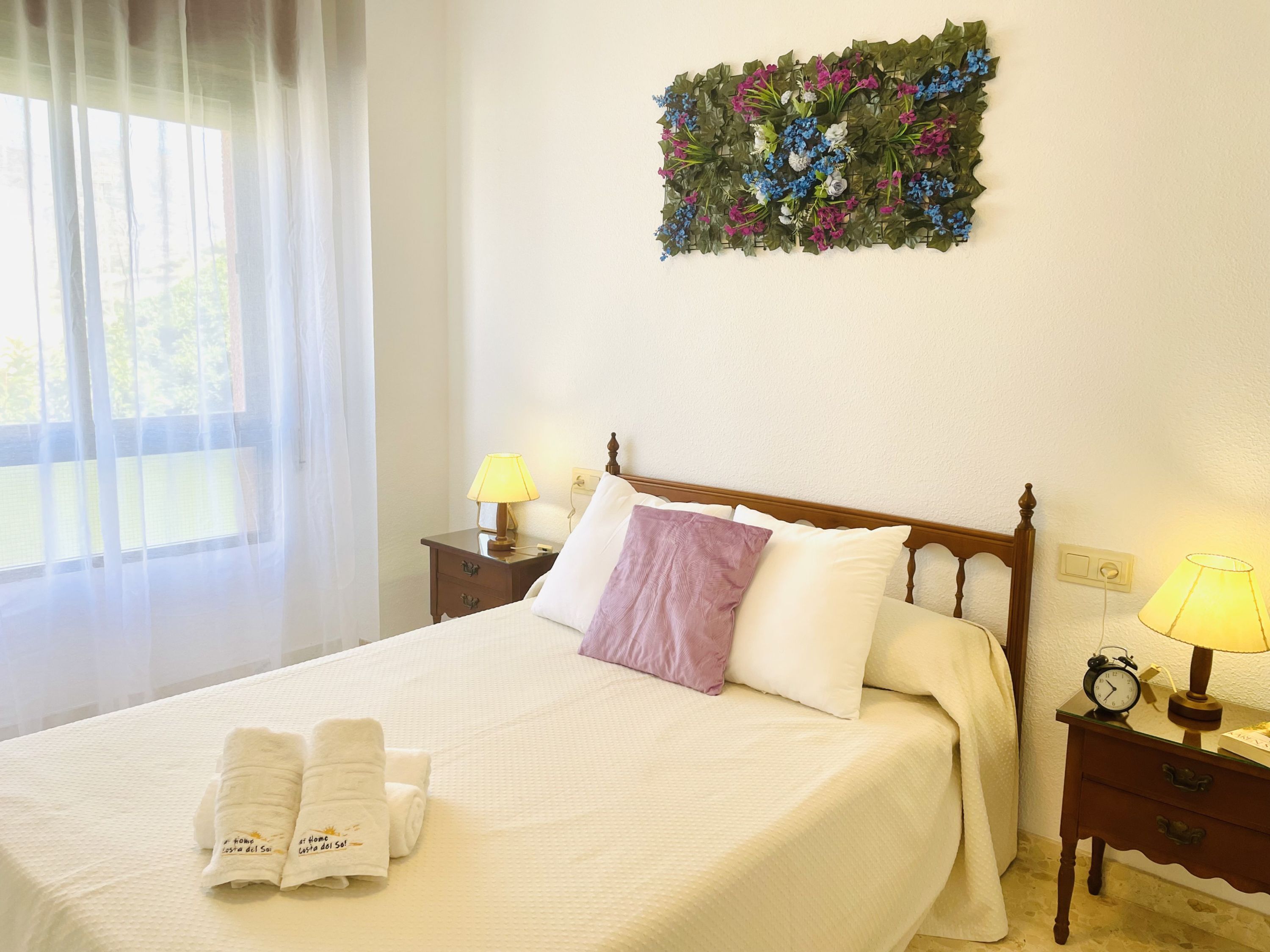 A neatly made bed with white linens, a purple pillow, and two towels with text. There is a wall-mounted floral decoration above the bed.