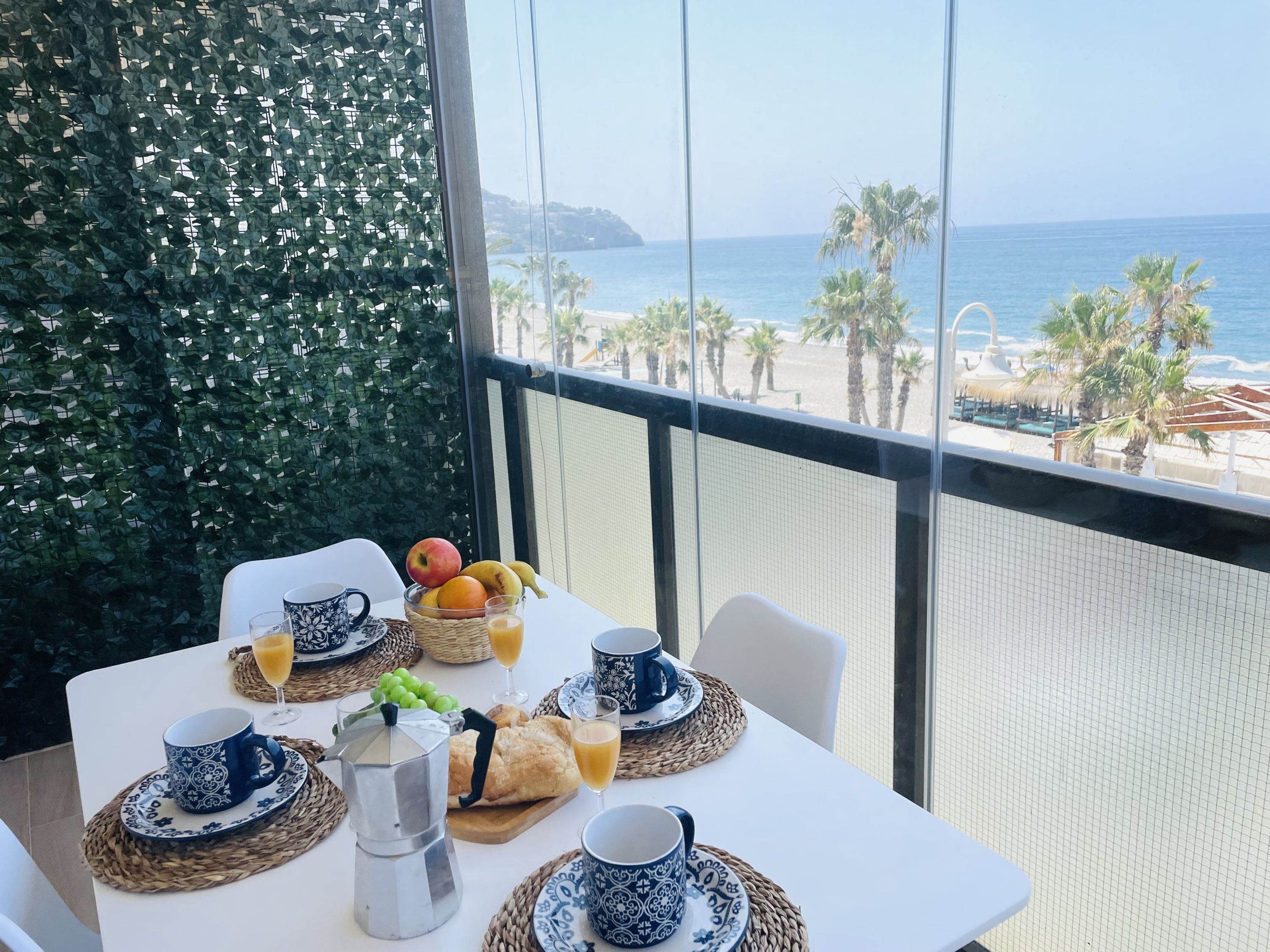 A balcony table with breakfast items, a view of the beach, and a decorative green wall.