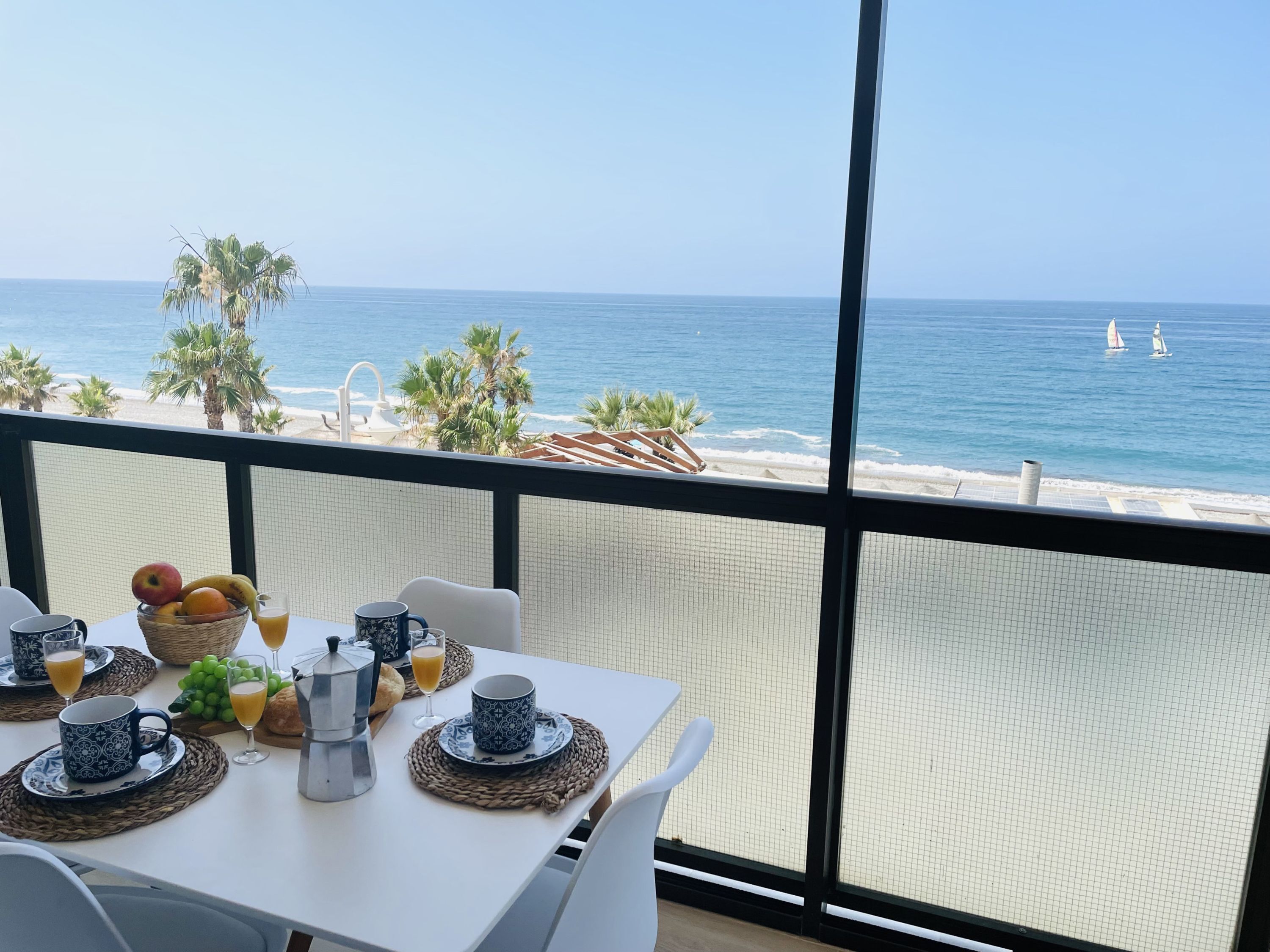 Varanda com mesa posta para o café da manhã, vista para uma praia com palmeiras e o oceano.