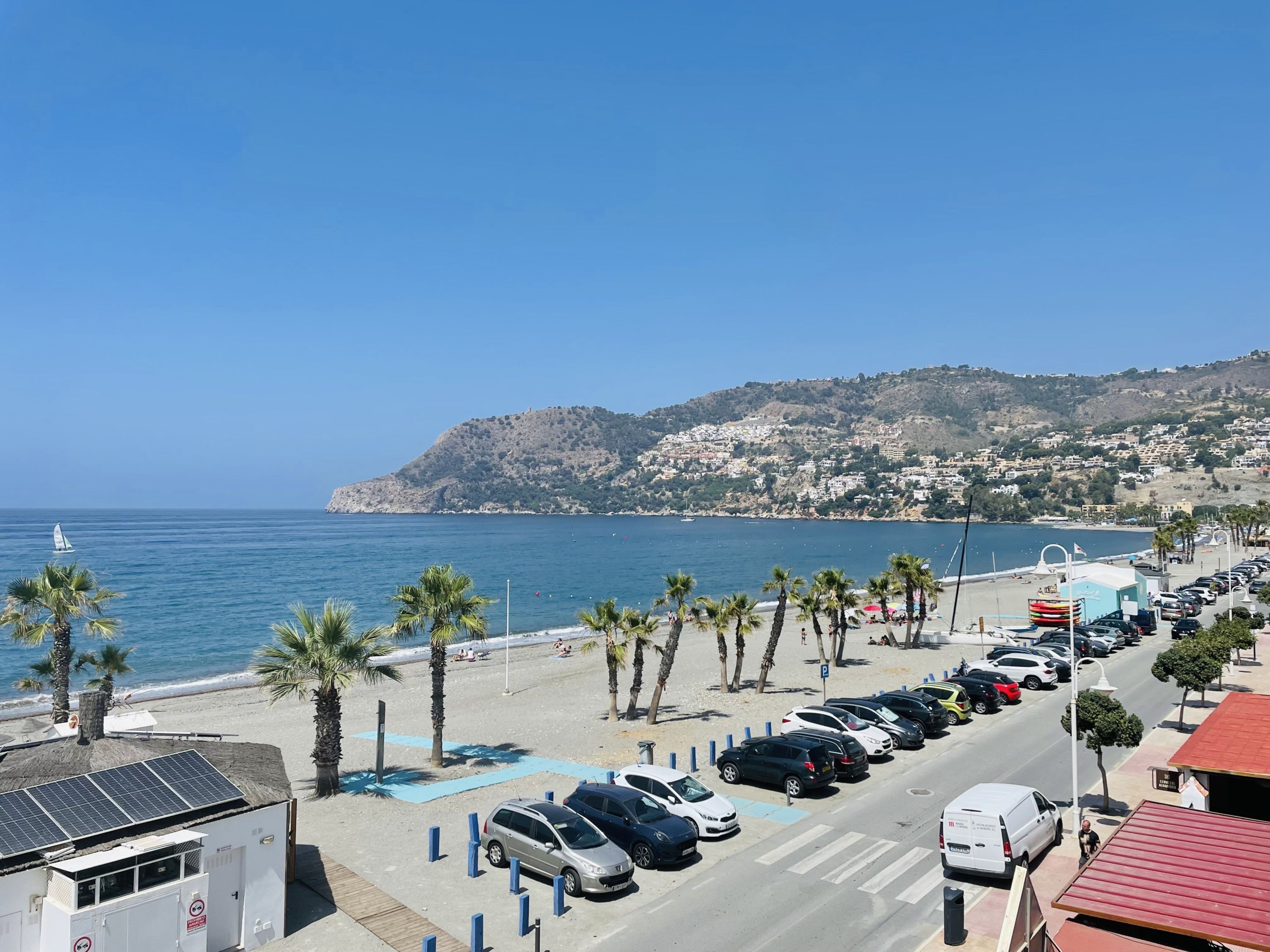 Una carretera costera con coches estacionados, una playa y un pueblo con colinas al fondo.