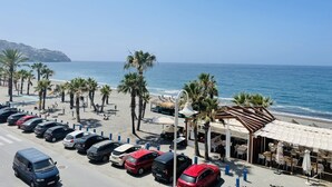 Un parking situé au bord de la plage, avec plusieurs voitures, des palmiers et un restaurant doté d’une terrasse offrant une vue sur l’océan.